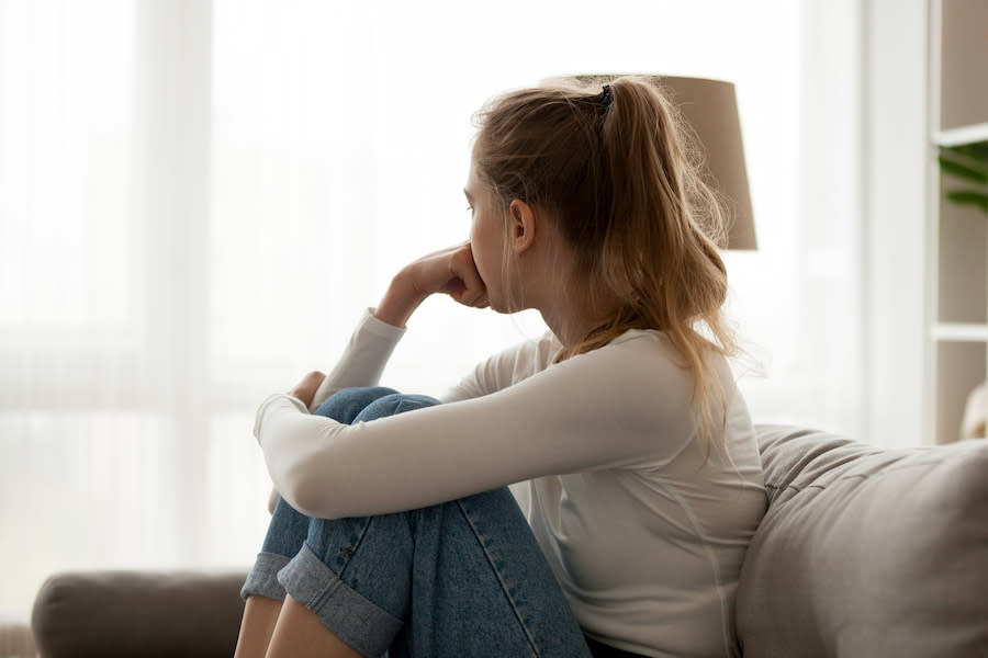 A young person sitting on couch looking away from camera with hand on chin and other arm hugging knees in.