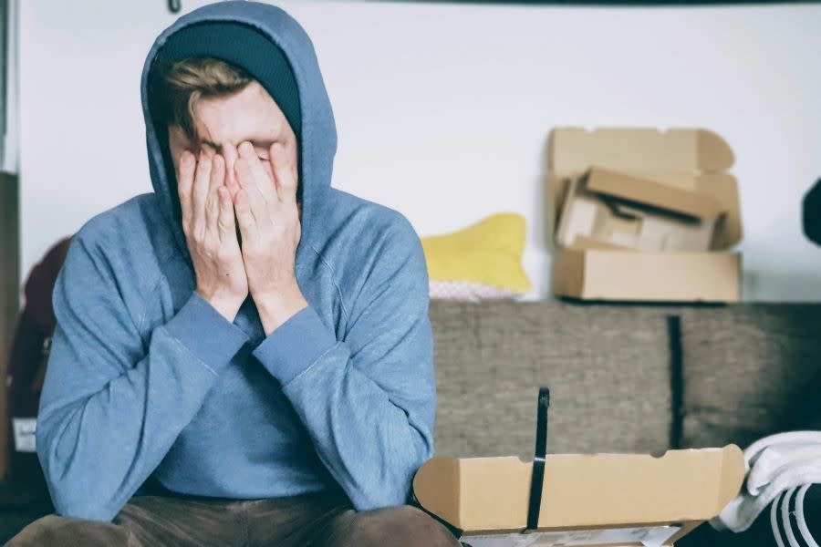 teenager sitting on couch in blue hoodie with hands covering face rubbing eyes.