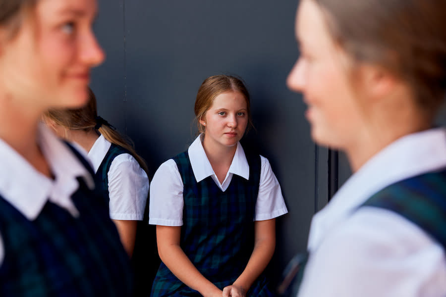 Image of a young teen girl sitting against a wall at school. She's looking at two other teen girls in the foreground, who are talking and laughing.