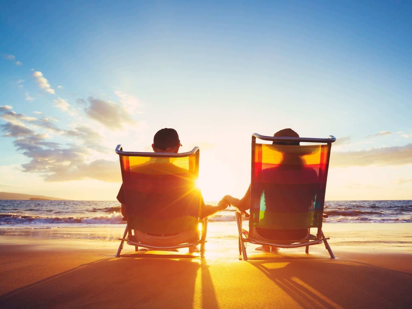 An old couple sitting together on a beach at sunset.