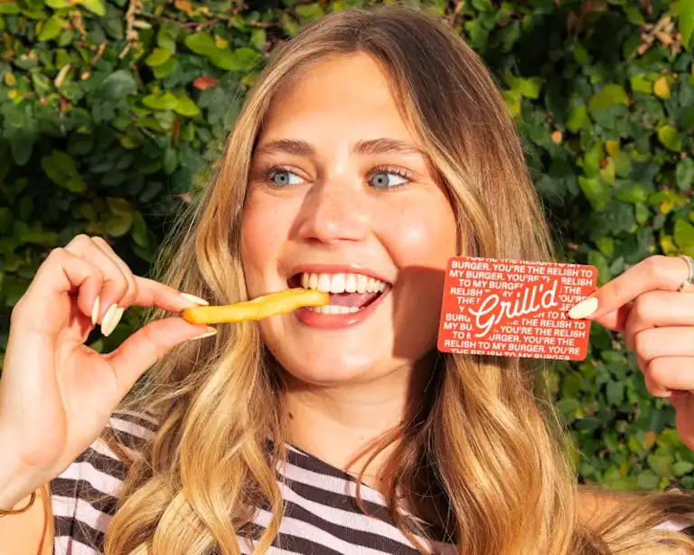 A smiling person outdoors holding a Grill’d gift card in one hand and a chip near their mouth with the other, with green foliage in the background.