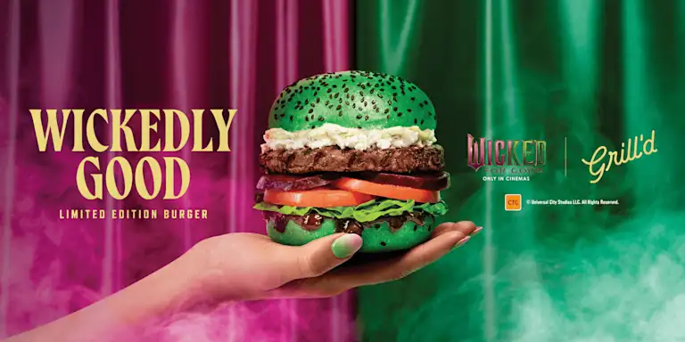 A hand with green painted nails presents a wicked-themed burger with a bright green bun sprinkled with black sesame seeds. The burger features layers of lettuce, tomato, beetroot, a beef patty, and creamy goats cheese and mayo. Behind it are dramatic pink and green smoky curtains with bold yellow text reading “Wickedly Good – Limited Edition Burger,” alongside the Wicked movie and Grill’d logos.