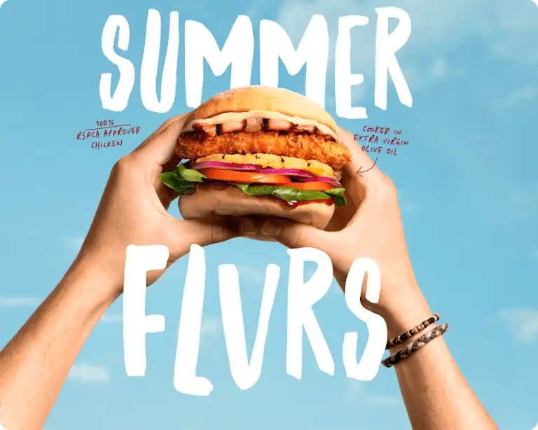 Two hands hold up a crispy chicken burger against a bright blue sky. The burger features a golden fried chicken fillet, bacon, pineapple, tomato, red onion, lettuce, and sauce on a soft bun. Text reads “SUMMER FLVRS” in bold white lettering.