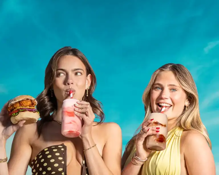 Two young women against a blue backgroun. One woman drinks a strawberry milkshake and holds a fried chicken burger, and the other woman is drinking a chocolate milkshake.