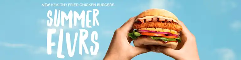 Two hands hold up a crispy chicken burger against a bright blue sky. The burger features a golden fried chicken fillet, bacon, pineapple, tomato, red onion, lettuce, and sauce on a soft bun. Text on the left reads “NEW HEALTHY FRIED CHICKEN BURGERS” and “SUMMER FLVRS” in bold white lettering.