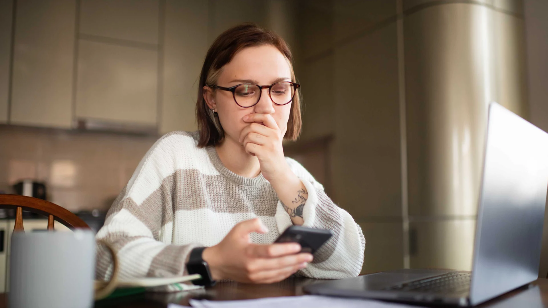 Voici comment vous protéger du spoofing Une jeune femme est assise dans la cuisine et regarde son smartphone avec inquiétude