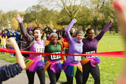 Enthusiastic female runners in tutus crossing charity run finish line in park, celebrating 