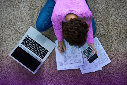 一个女人坐在地板上处理税务，手里拿着笔记本电脑和计算器