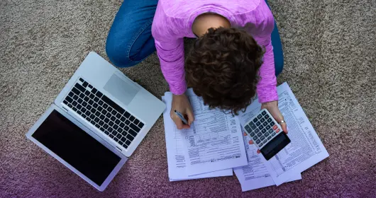 一个女人坐在地板上处理税务，手里拿着笔记本电脑和计算器