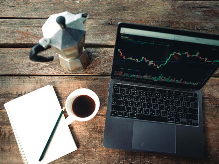Laptop mit Aktienmarktgrafiken neben einer Kaffeetasse, einer Espressomaschine und einem Notizbuch auf einem Schreibtisch