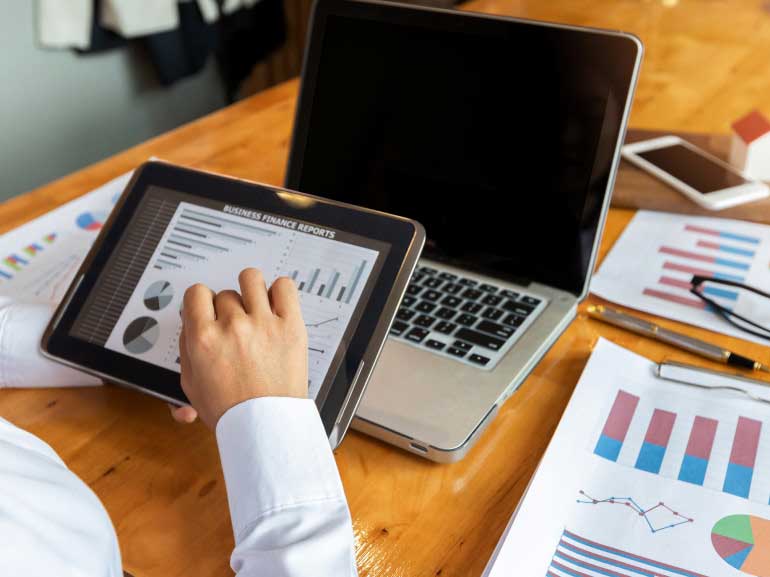 A businessman studying charts on his tablet in front of his laptop and reports on the desk