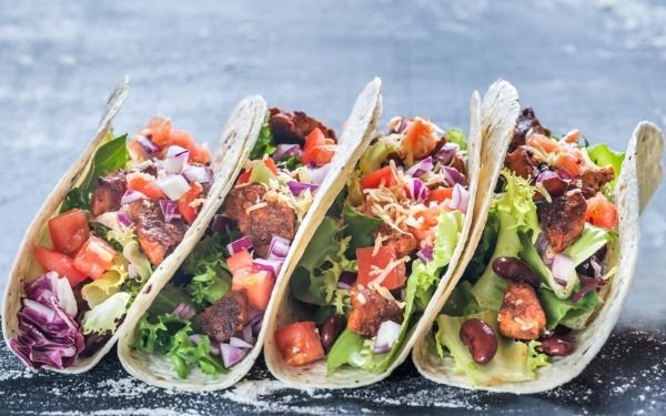 Four tacos with pico de gallo, lettuce, beans, meat and cheese lined up leaning on each other on a table.