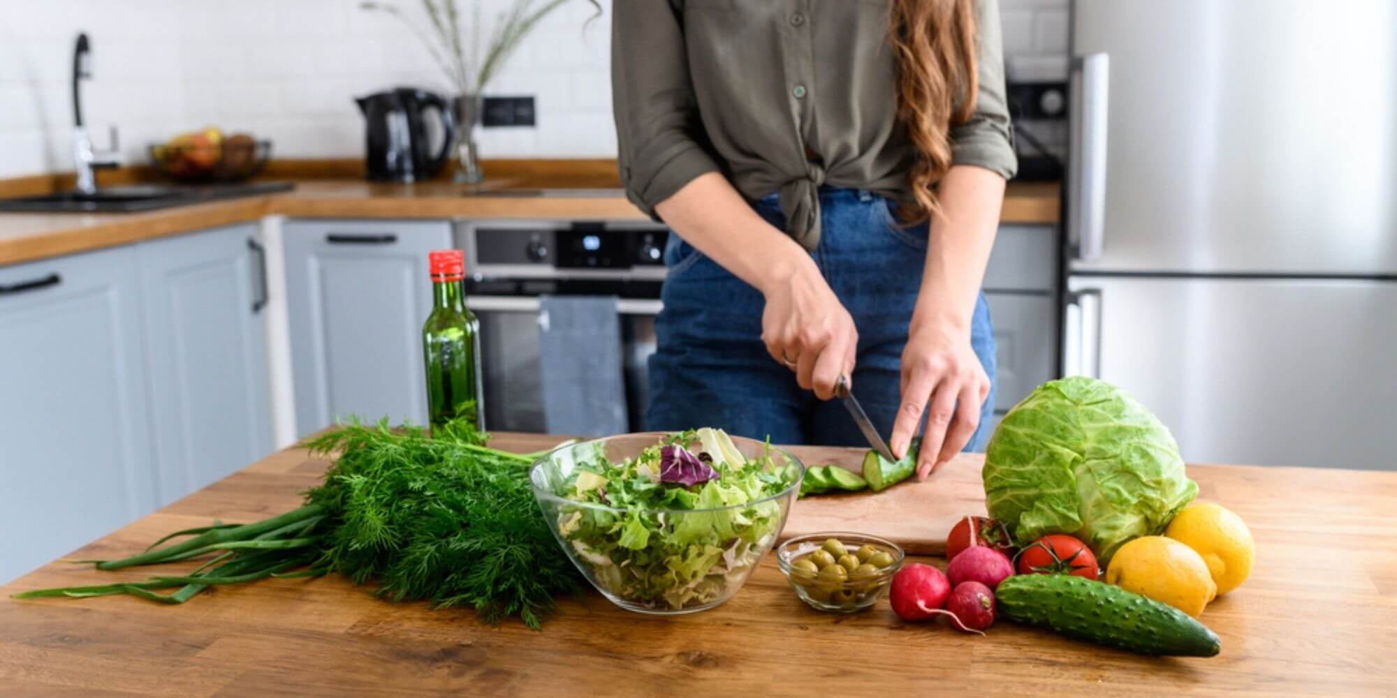 Woman slicing cucumbers on a wooden cutting board in a kitchen next to lettuce, olives, tomatoes, and lemons.