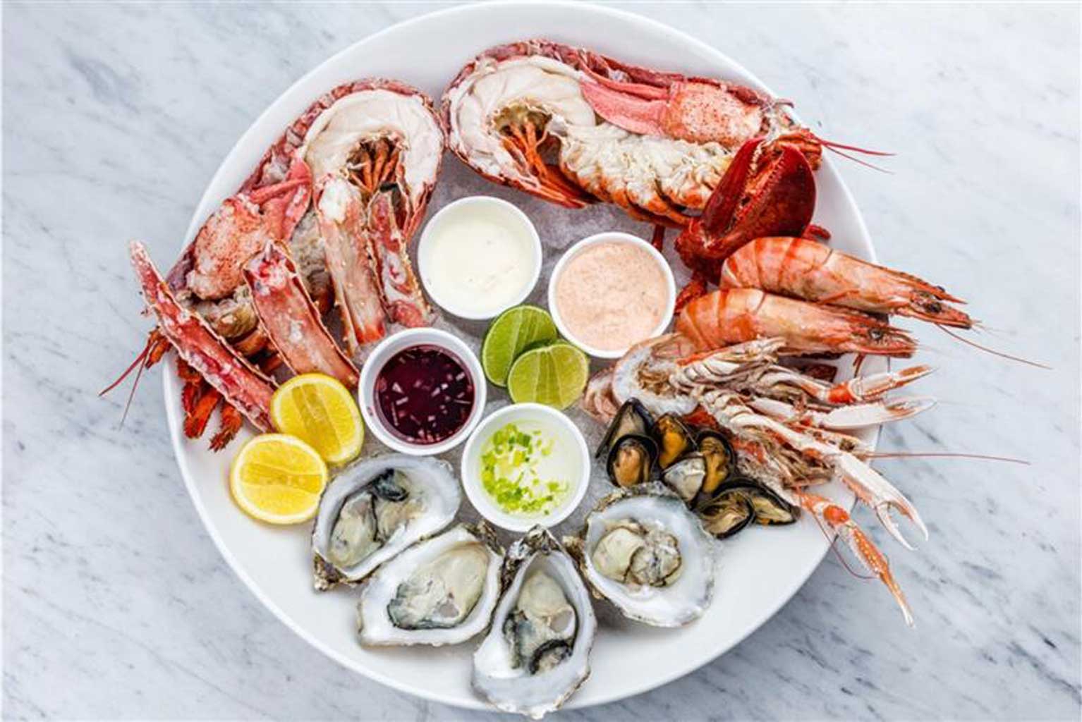 a fresh seafood plate full of oysters, shellfish, lobster, and mussels over ice with garnishes of lemon, lime and assorted dipping sauces