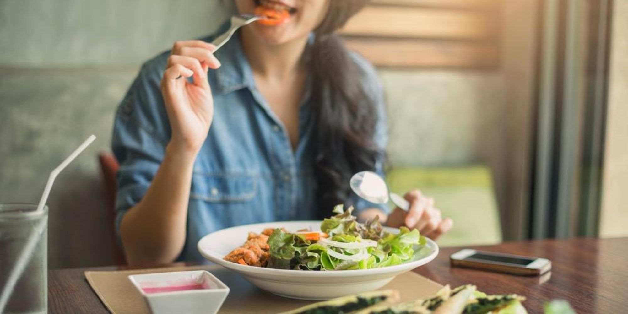 Woman with dark brown hair eating a salad with a fork and spoon in her hands
