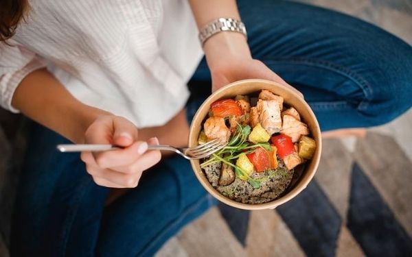 woman eating a bowl of salmon, bell peppers, and quinoa in a bowl with a fork in her hand
