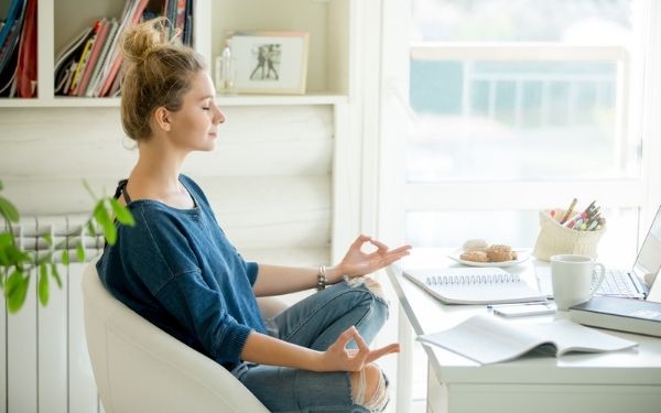 Woman meditating on white chair next to desk with a computer, coffee cup, and work papers.