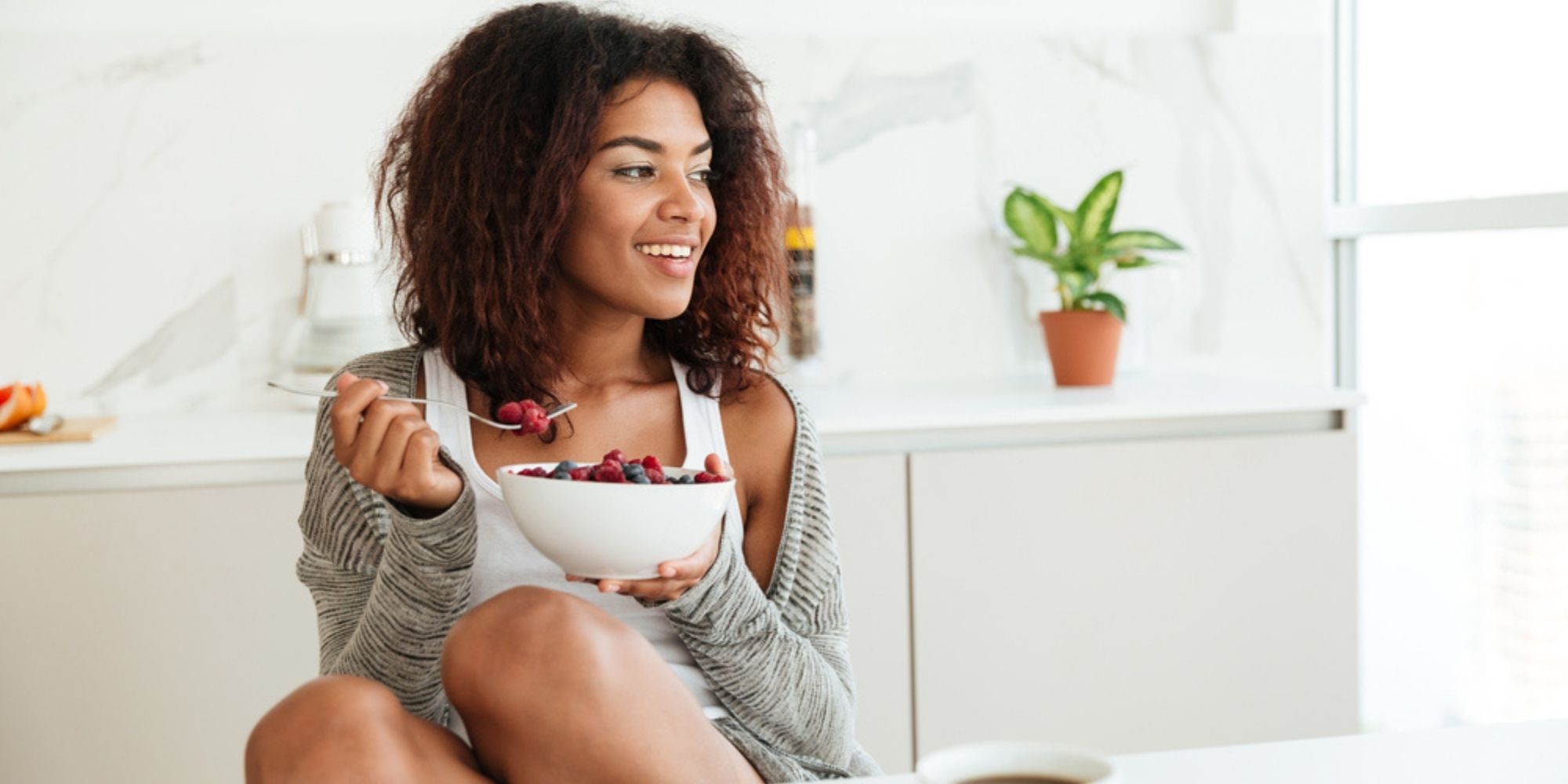 Woman with curly hair sitting at a white table eating a bowl of raspberries, strawberries, and blueberries