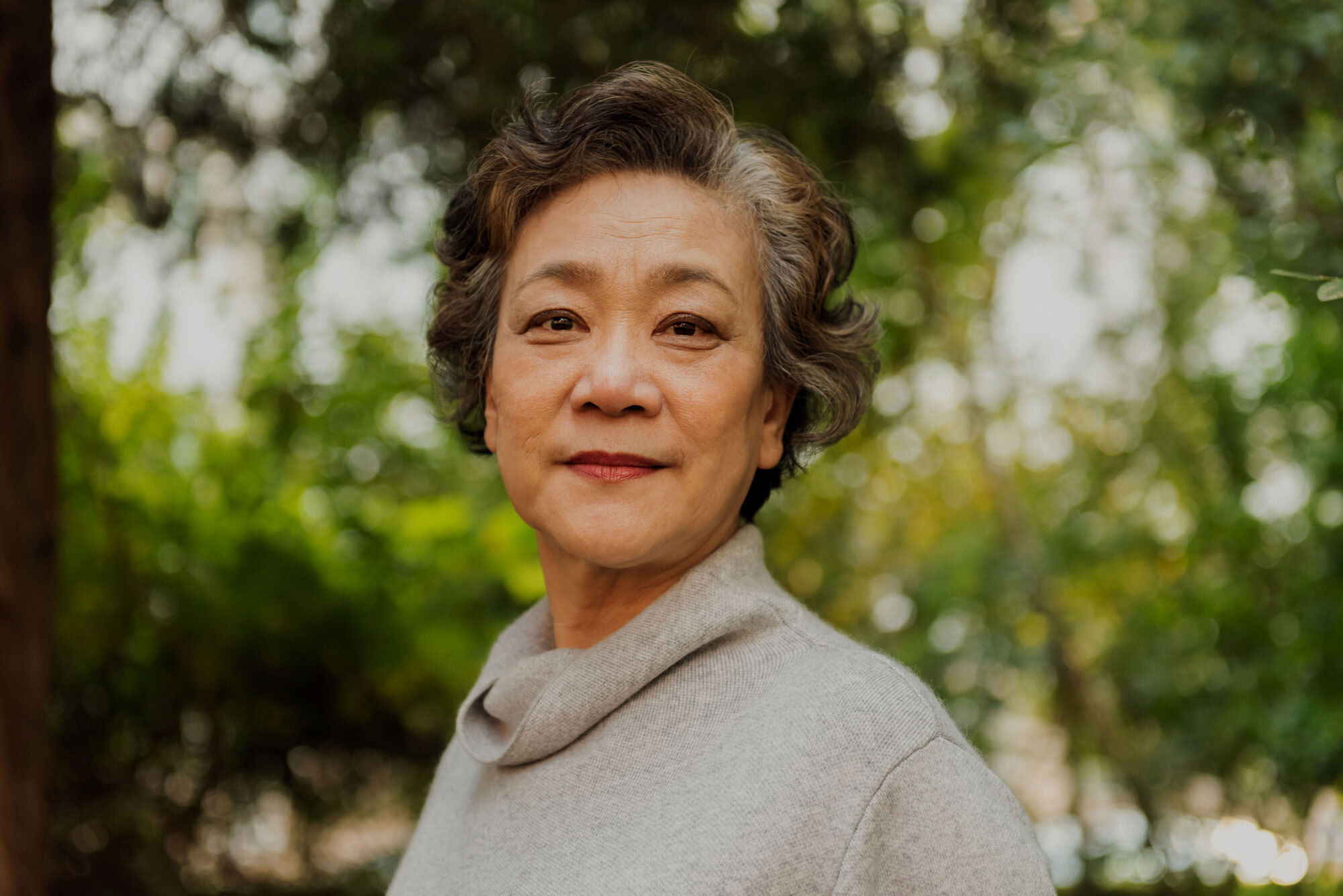 An older asian woman in a gray sweater standing in front of blurred out trees.