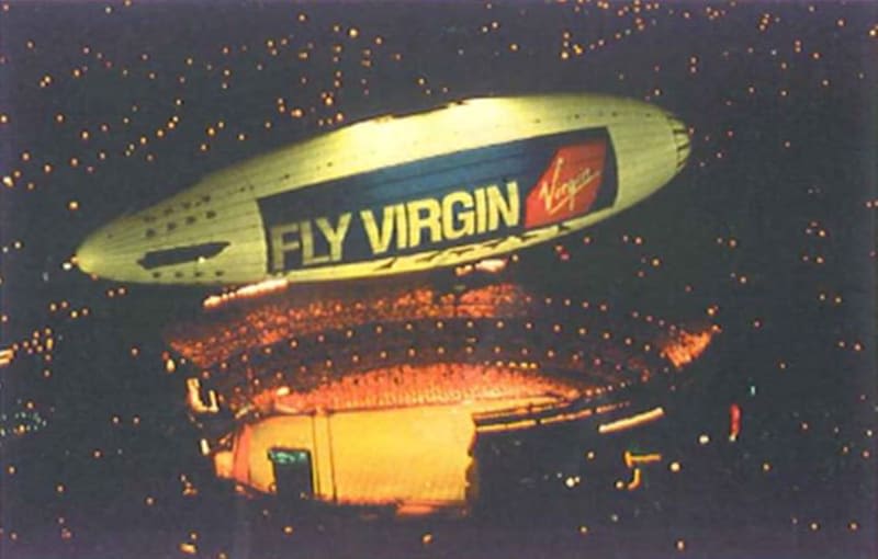 An airship with the text Fly Virgin and the Virgin logo on it flying above a stadium
