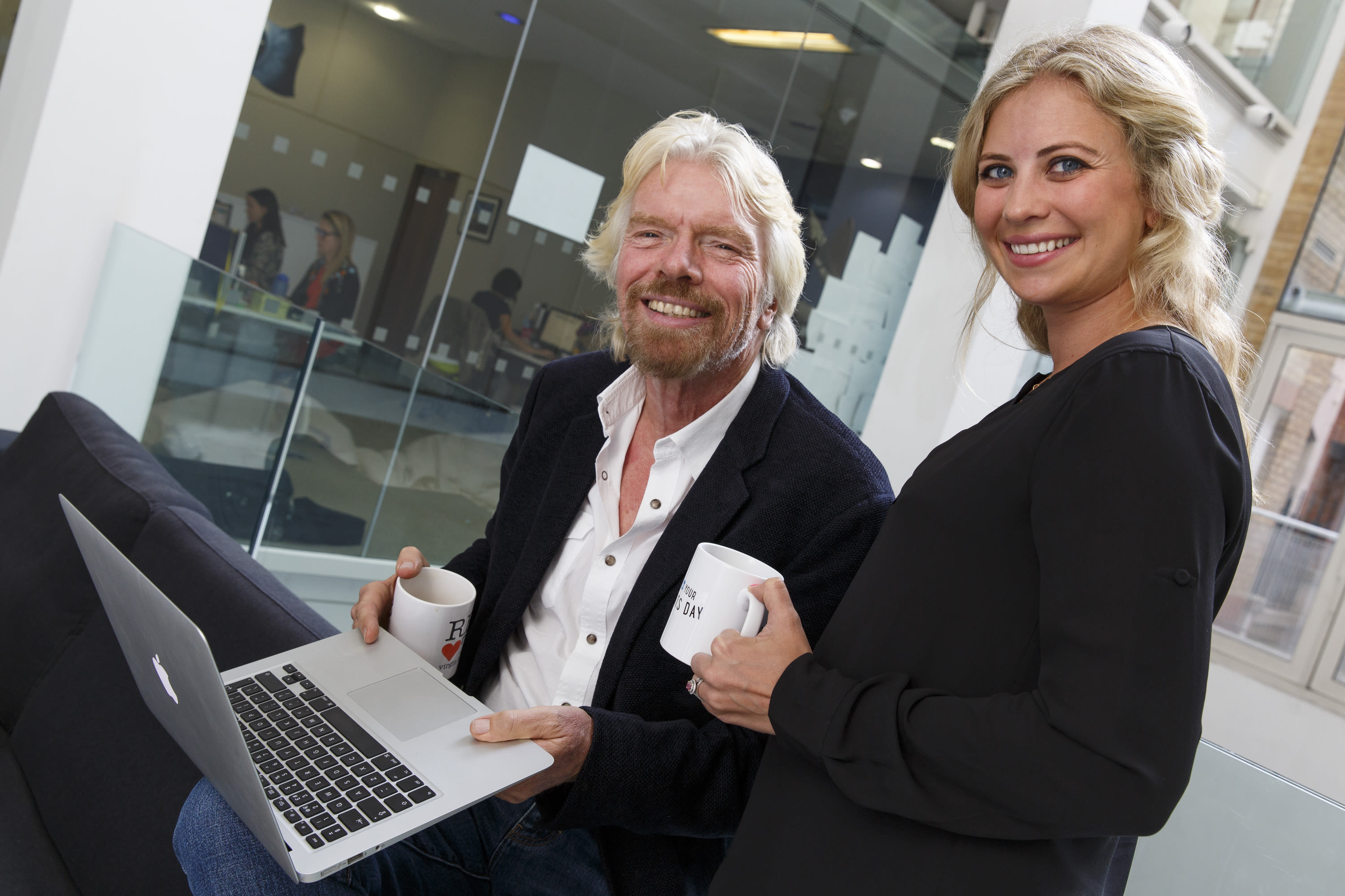 Richard Branson and Holly Branson smiling in an office and holding a mug and laptop