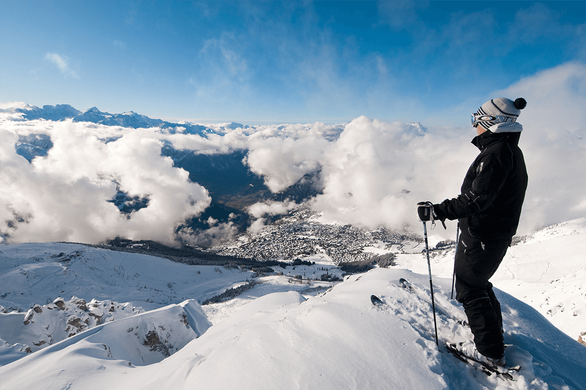 A skier stands on the edge of a slope, looking at the mountains and clouds below