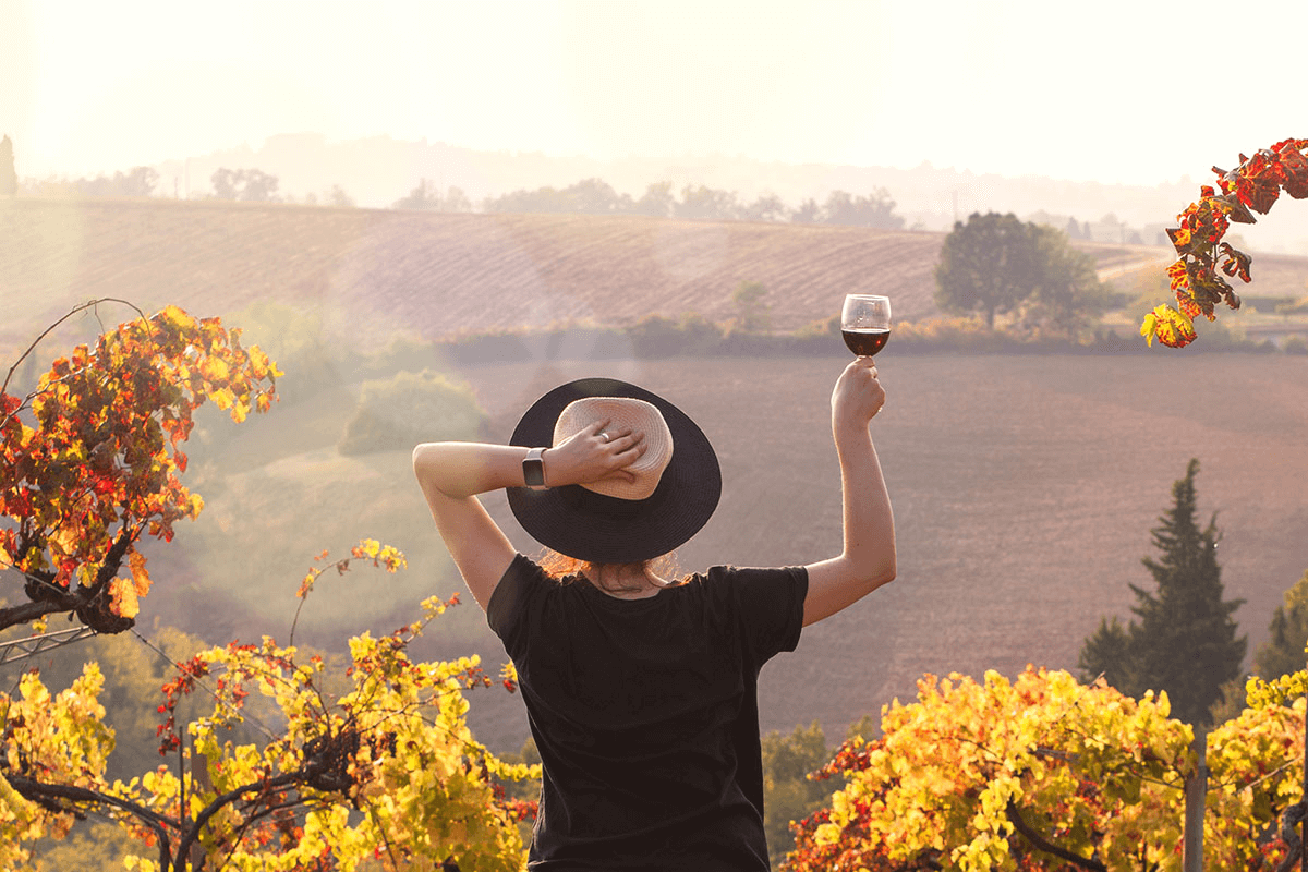Woman in a hat holds up a glass of red wine and looks out over a winery