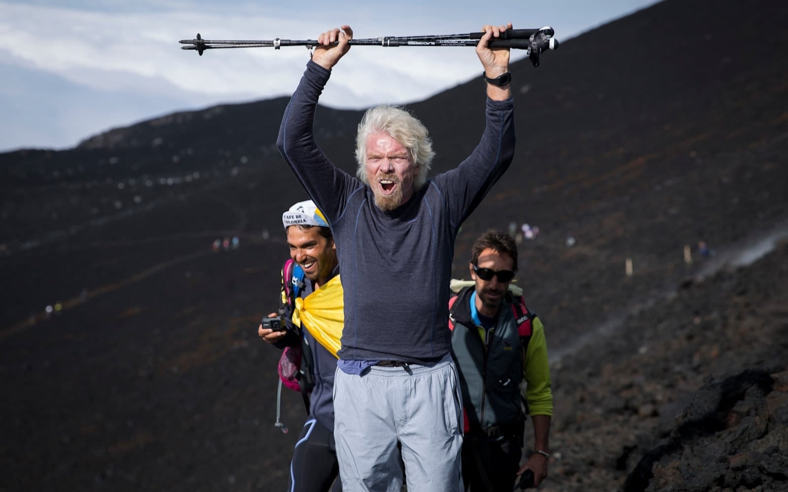 Richard Branson on a hike cheering and lifting walking poles above his head. There are two people behind him and mountains in the background