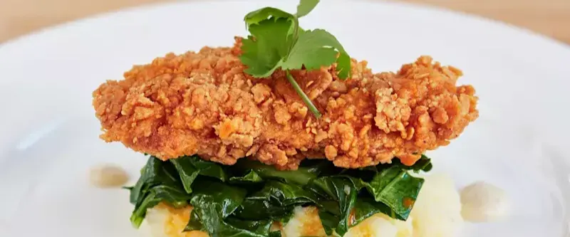 Synthetic meat tender with a piece of herb sitting on top as it rests on spinach, all on a white plate 