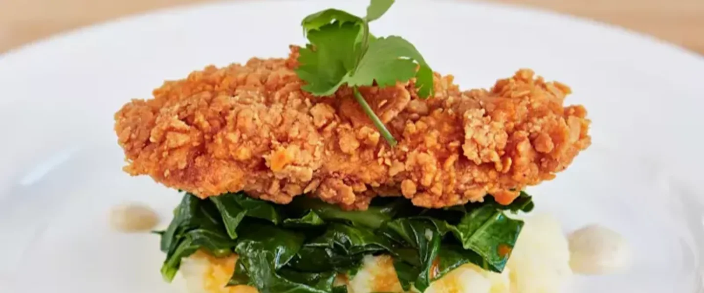 A piece of fried chicken on a bed of spinach and mash potatoes on a white plate.