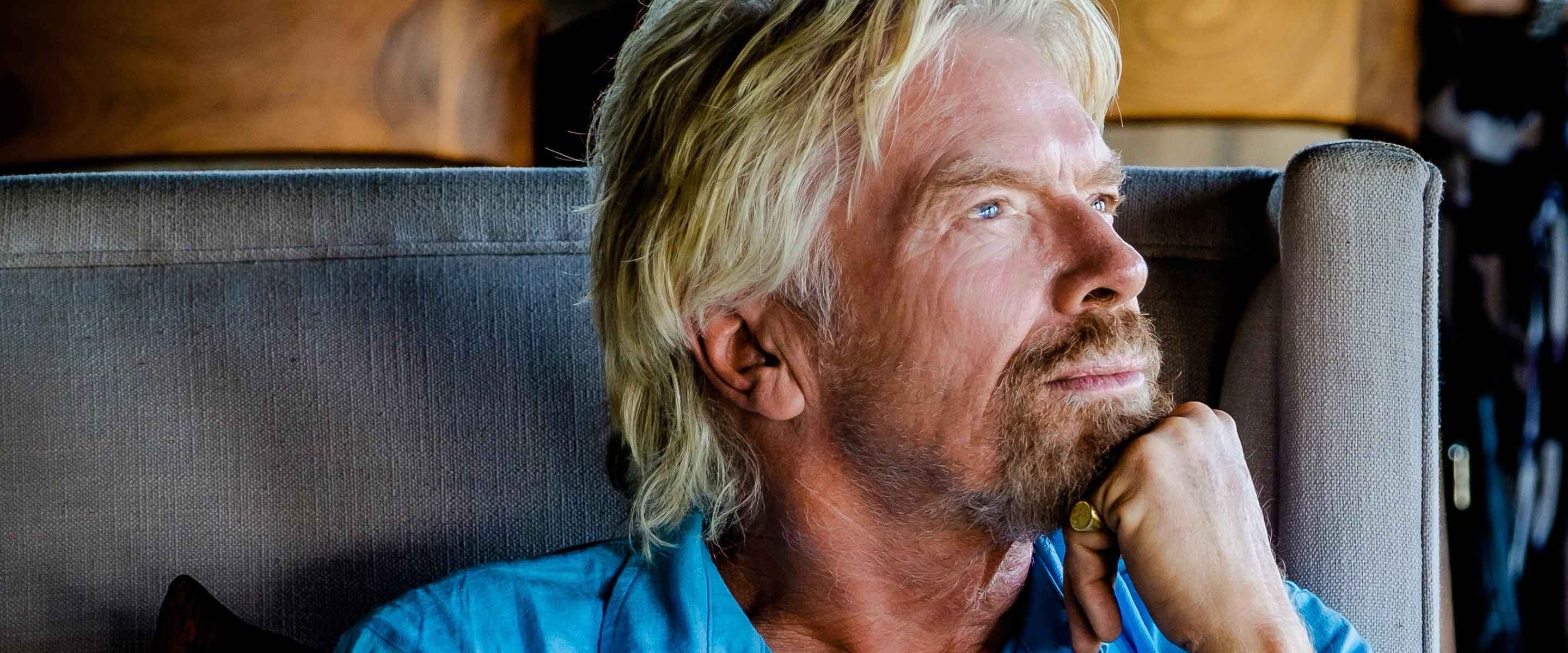 Sir Richard Branson sits in an armchair looking deep in thought with his chin resting on his hand. He is wearing a light blue, casual button down shirt