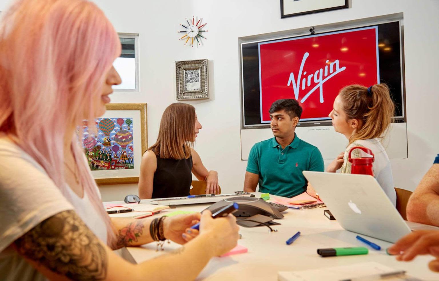 People sitting around a table talking, a screen in the background has the Virgin logo on it