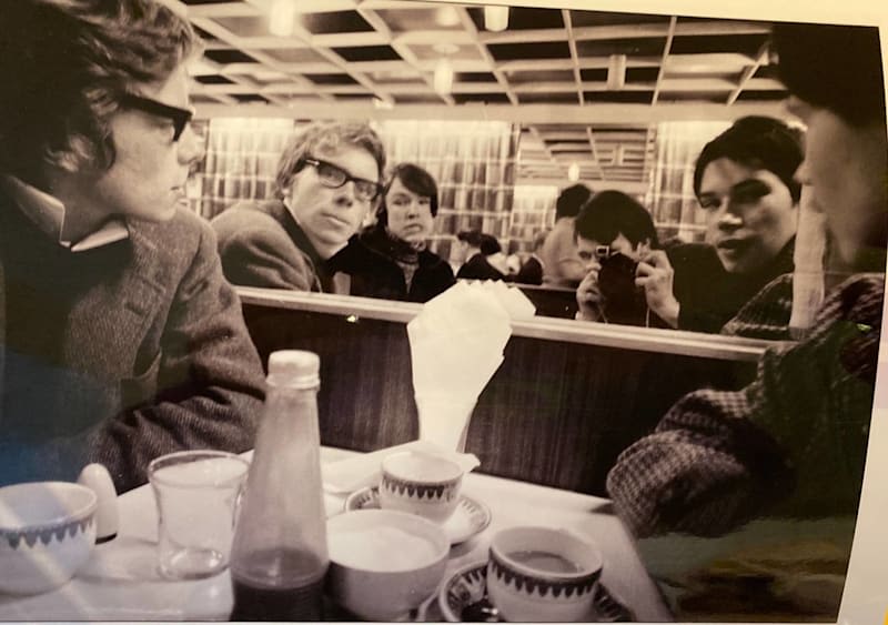 Richard Branson as a young man sits in a restaurant with his friends including Nik Powell