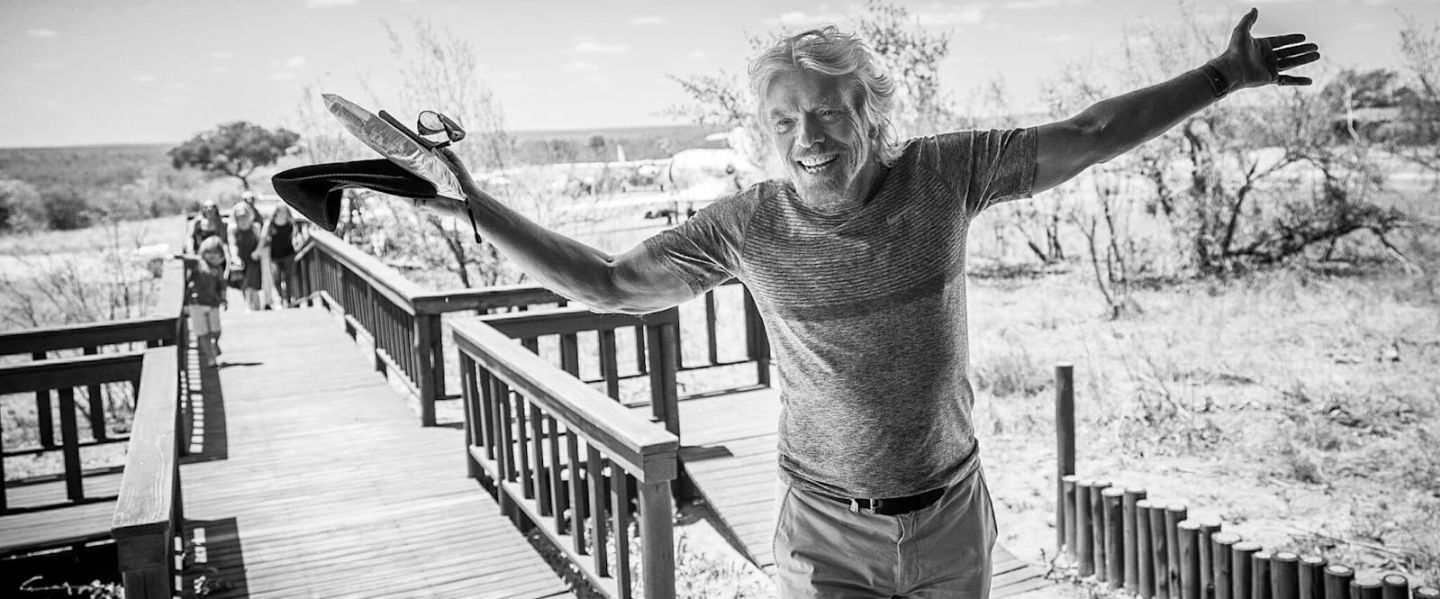 Black and white photo of a smiling Richard Branson with his arms wide open on a wooden walkway in open grassland
