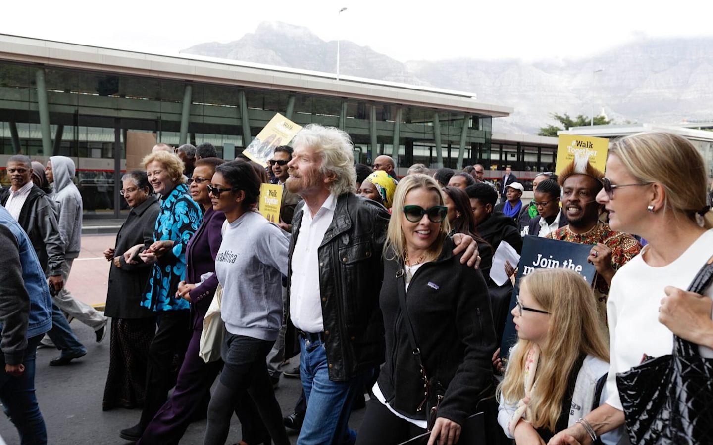 Richard Branson walks with a large group of people. He is wearing jeans, a black leather jacket and a white shirt