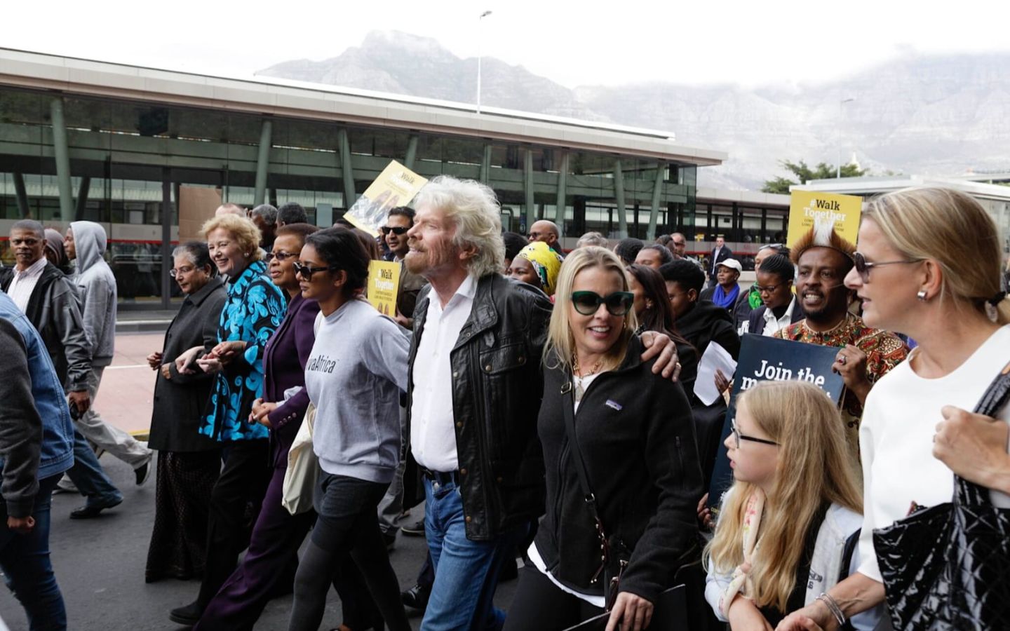 Richard Branson walks with a large group  of people.  He is wearing jeans, a black leather jacket and a white shirt