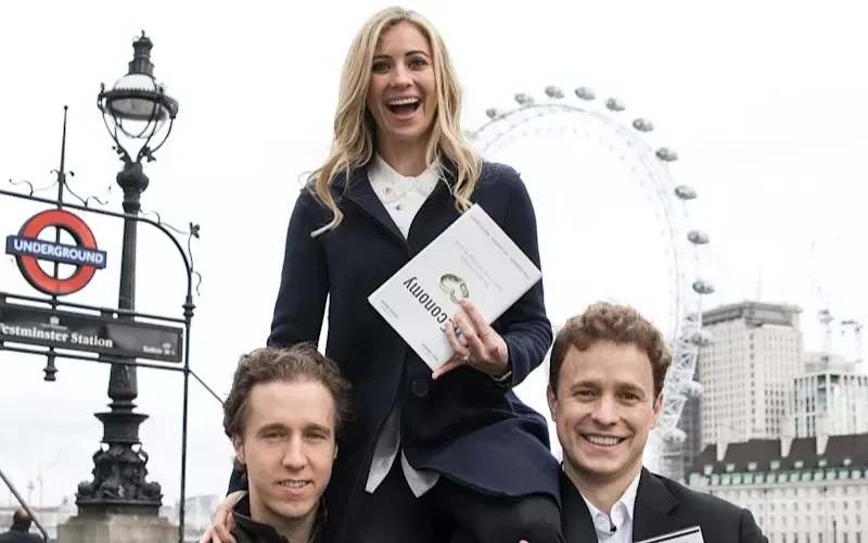 Holly Branson sitting on the shoulders of Craig and Marc Kielburger holding a copy of Weconomy in front of the London Eye 