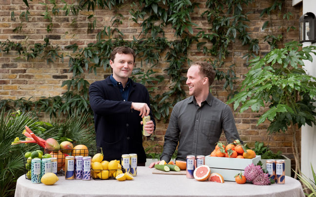 Jack Scott and Alex Wright, co-founders of Dash Water, stand behind a table that has fruits including lemons, grapefruits and rhubarb, and colourful cans of Dash Water on it