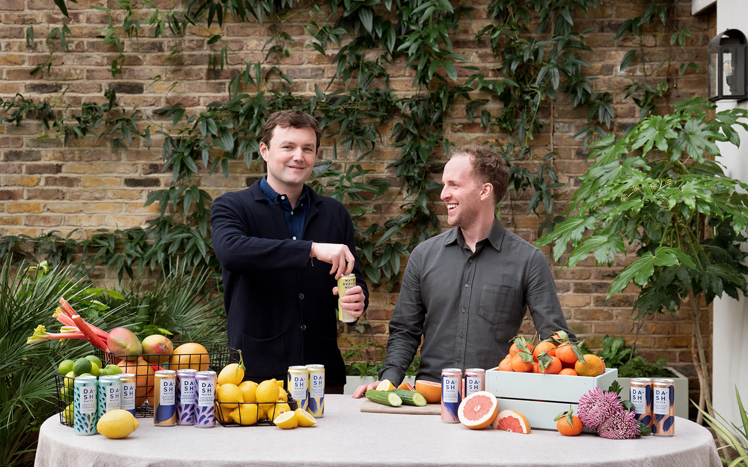 Jack Scott and Alex Wright, co-founders of Dash Water, stand behind a table that has fruits including lemons, grapefruits and rhubarb, and colourful cans of Dash Water on it