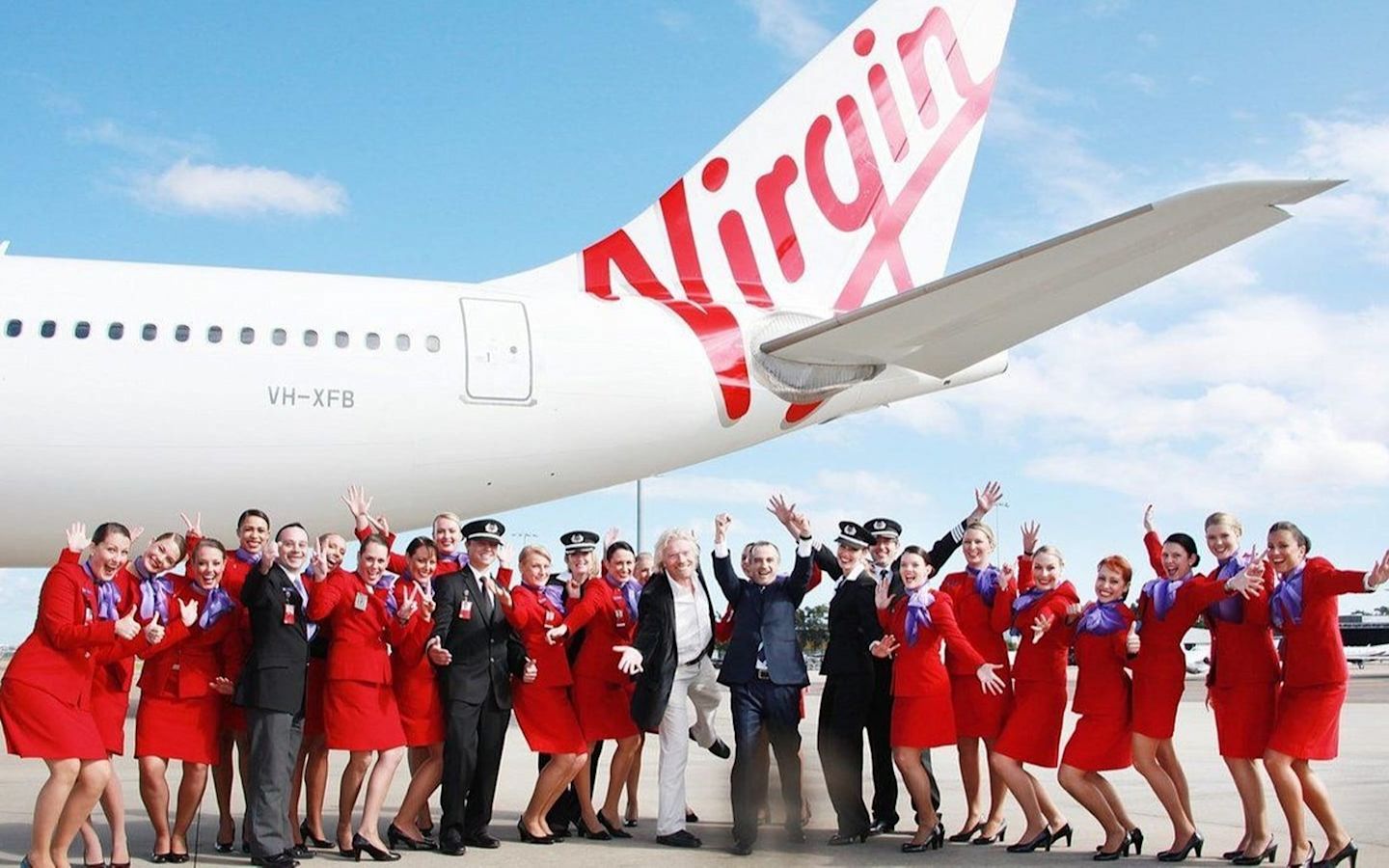 Richard Branson, Virgin Australia cabin crew and pilots stand with their arms in the air, smiling, in front of a Virgin Australia plane