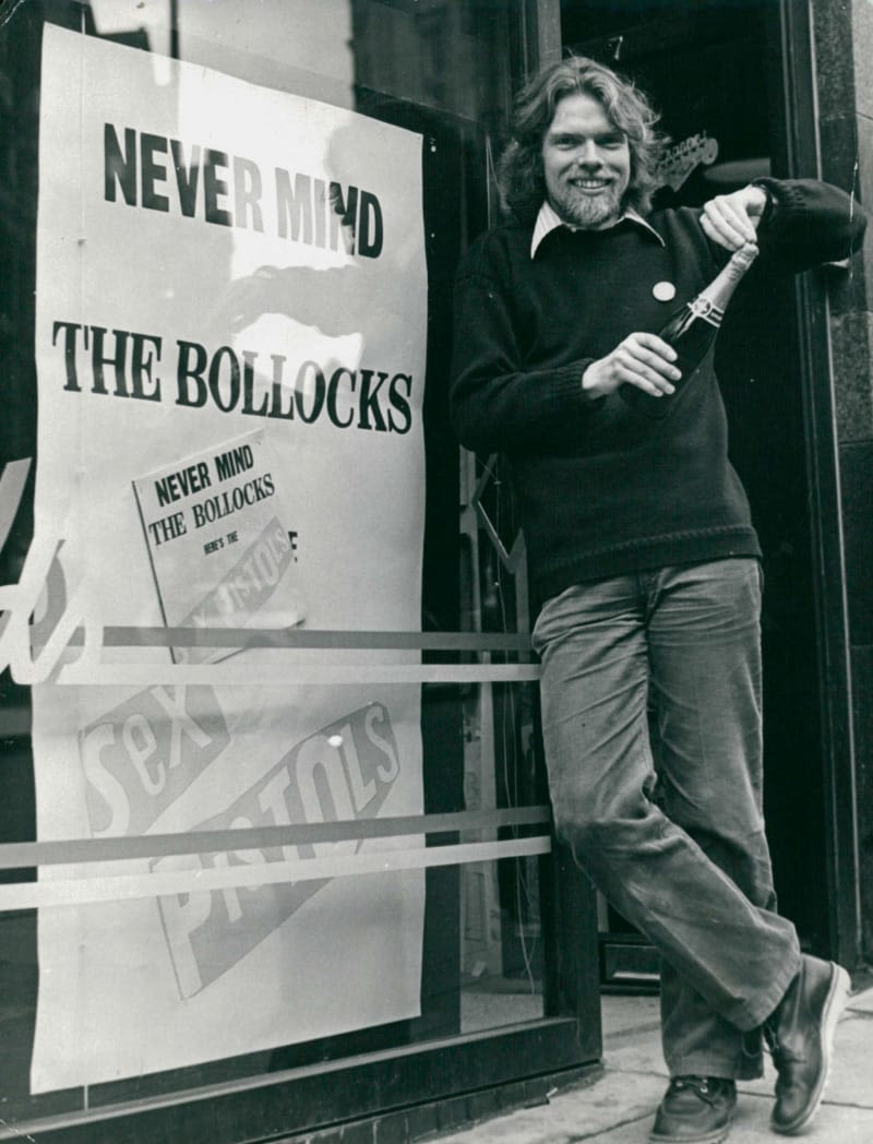 Richard Branson holding a bottle of champagne in front of a sign that reads 'Never Mind The Bollocks'