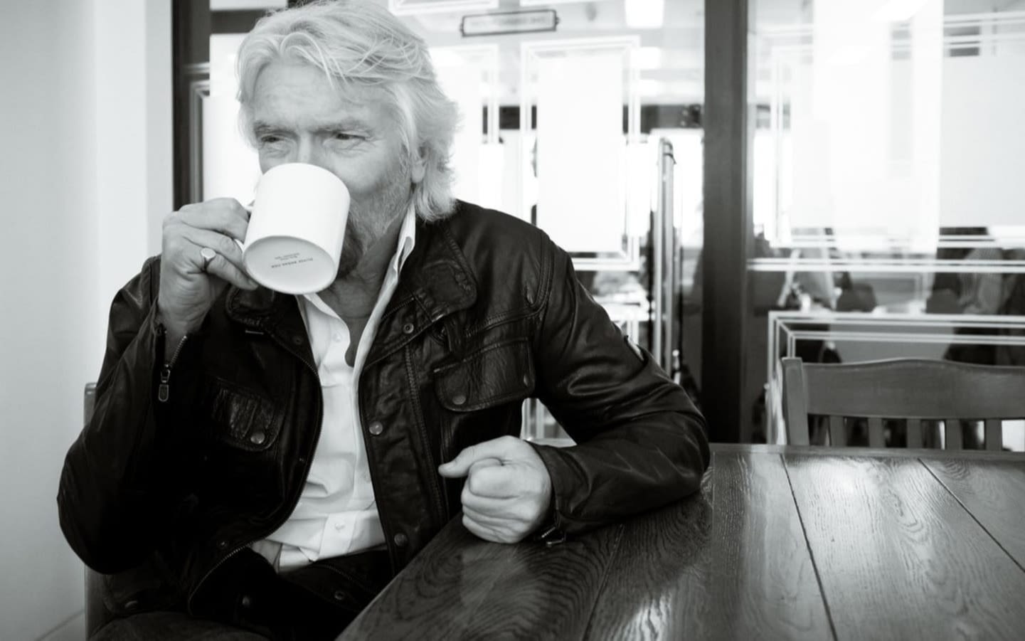 Black and white image of Richard Branson sitting at a table drinking from a mug