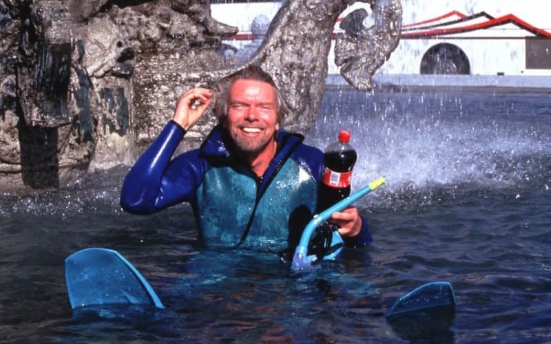Richard Branson, floats in a pool smiling, holding a bottle of Coca Cola and a snorkel in his arm.  He is wearing a blue wet suit, complete with fins.