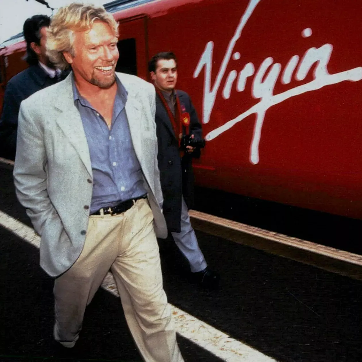 Richard Branson smiling with his hands in his pockets walking alongside a Virgin Train