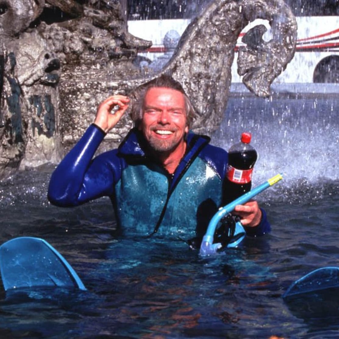 Richard Branson in a wet suit sitting in a fountain with a Virgin Cola bottle