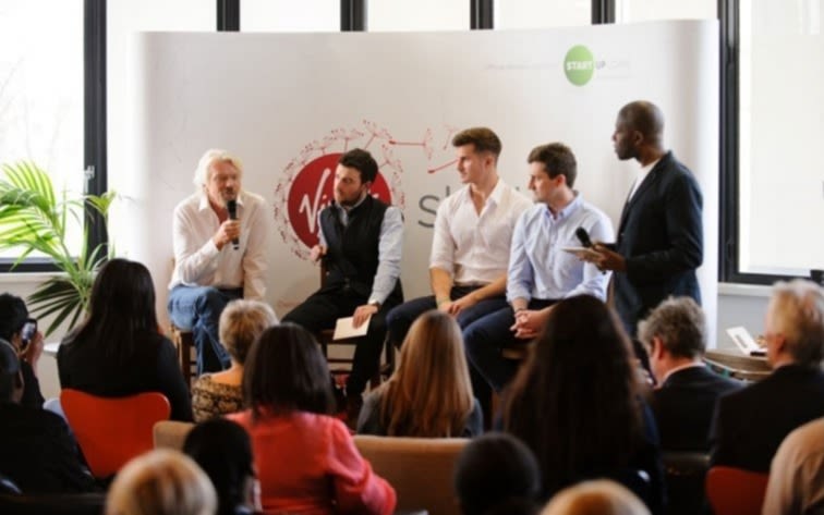 Richard Branson speaking into a microphone on a panel with 3 other men.