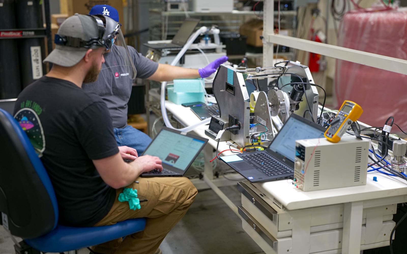 picture of 2 men sat at a work bench with 2 laptops both wearing visors look to be testing a ventilator in the process of being made