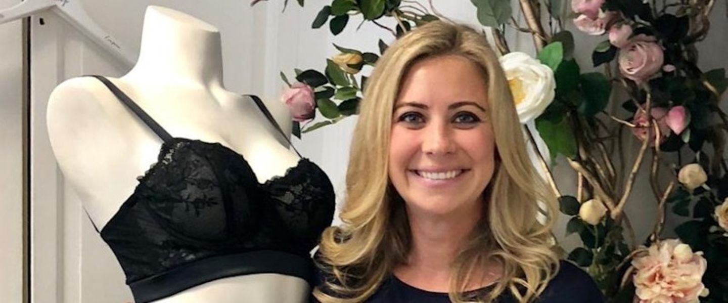 Holly Branson smiling at the camera, next to a mannequin wearing a black bra and in front of a wall with flowers climbing up it