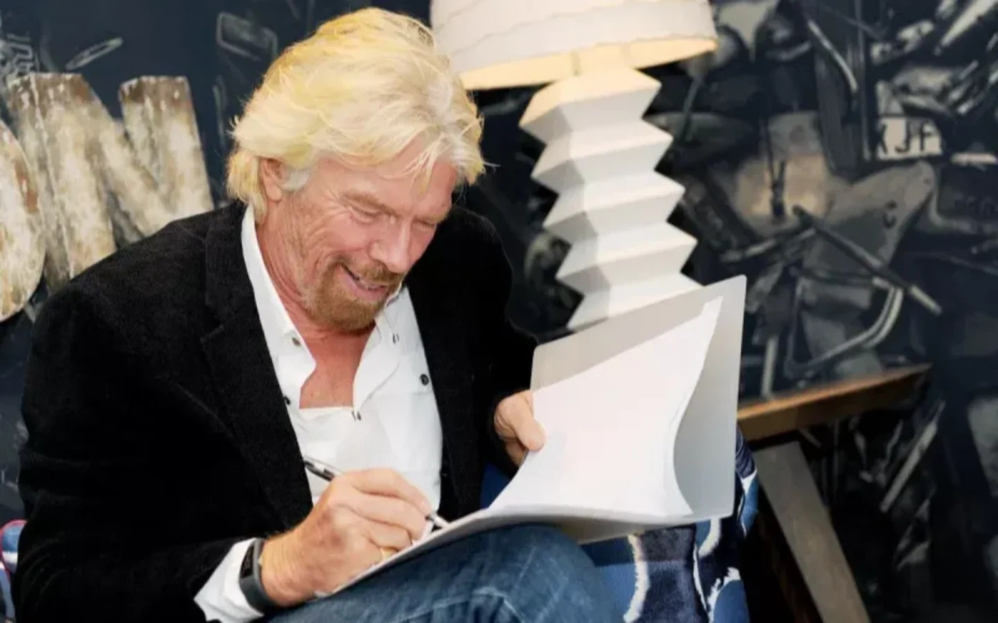 Richard Branson sitting in a chair writing and smiling