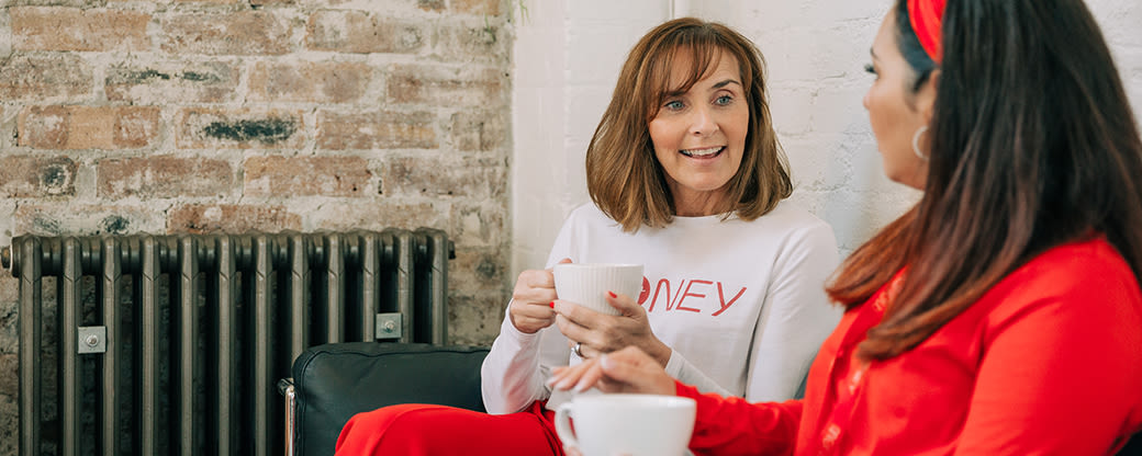 Two women having a cup of coffee and chatting, one is wearing a white top with the Virgin Money logo on